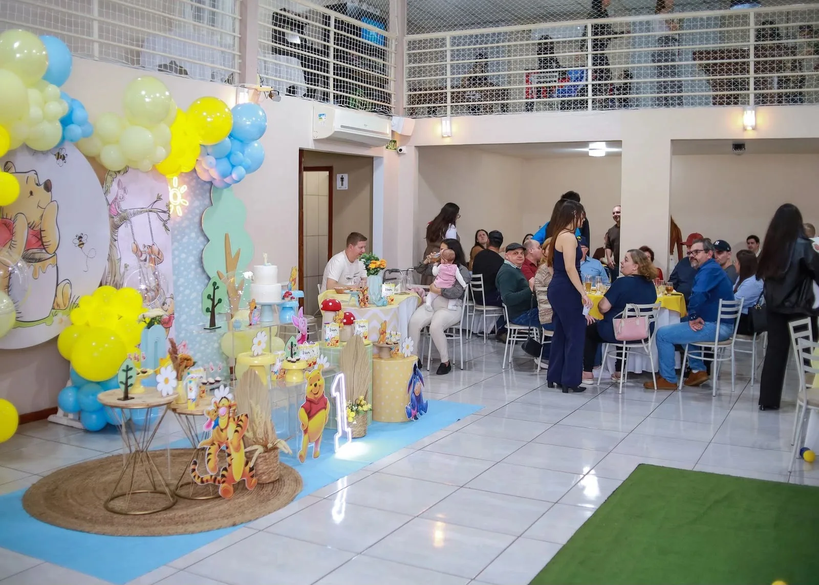 Mesa de festa de um ano no tema do Ursinho Pooh, com painel ilustrado, bolo e convidados ao fundo