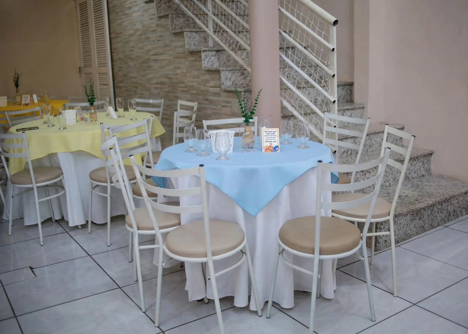 Mesas redondas postas com toalhas azul e amarela, taças e arranjos, ao lado da escada do salão