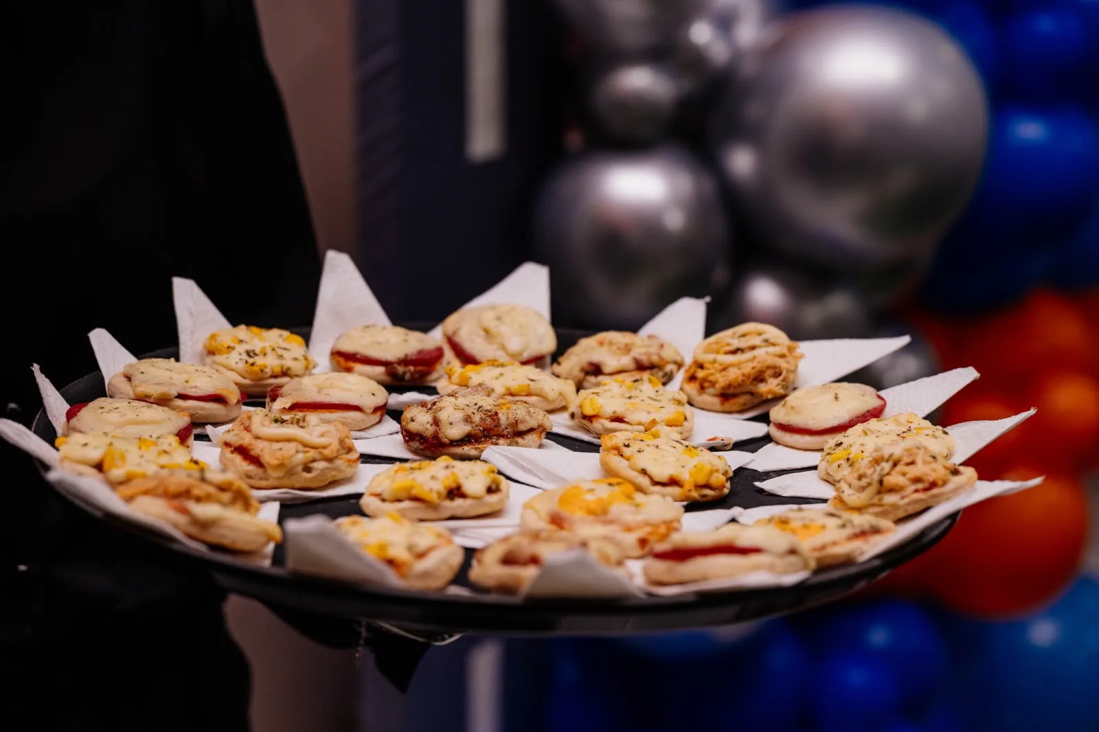 Bandeja redonda de mini pizzas servida durante a festa, com balões ao fundo