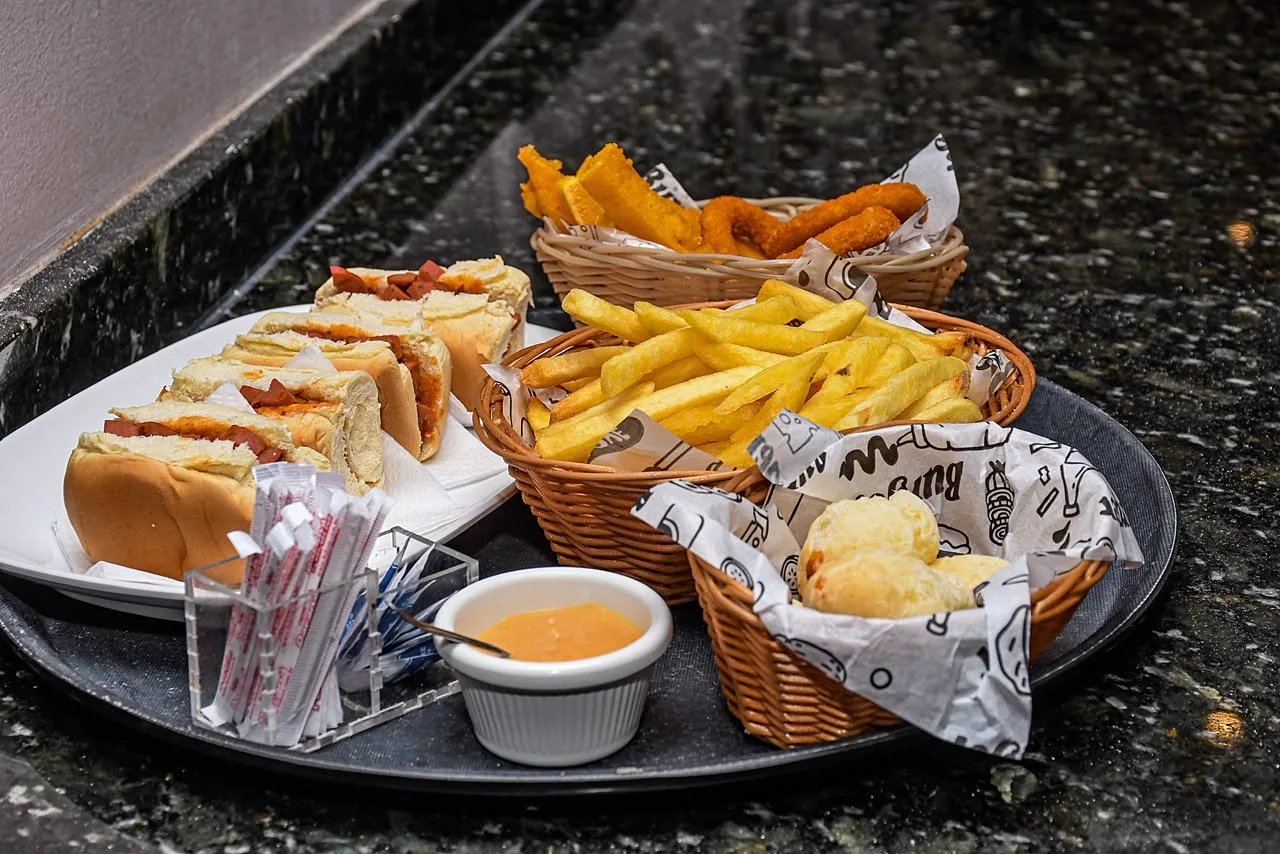 Bandeja com sanduíches de metro fatiados, cesto de batata frita, pão de queijo e molho